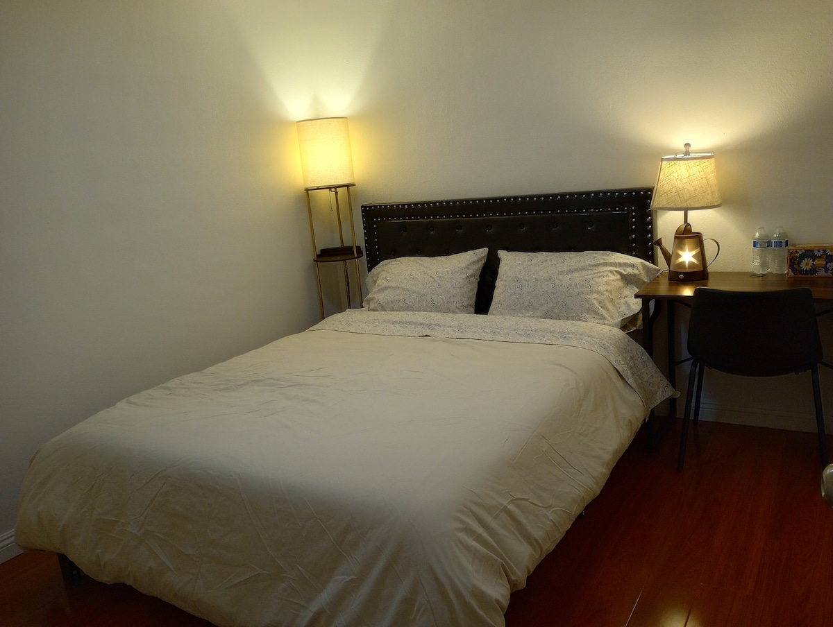 A cozy bedroom features a spacious bed with neutral bedding arranged neatly. Soft lighting is provided by two table lamps on either side, illuminating the room. A small desk and chair are positioned against the wall, enhancing the functional layout.