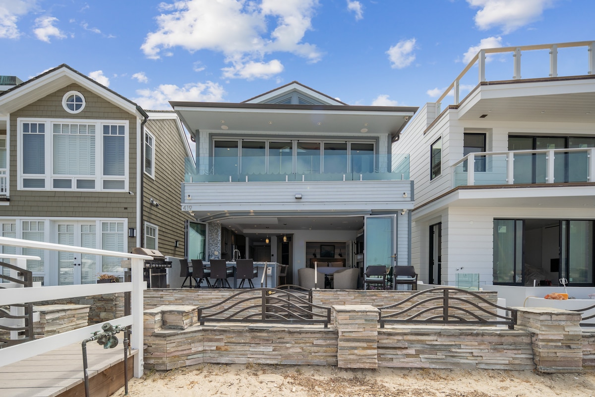 A modern beachfront property is displayed, featuring a spacious balcony and large glass doors that open to the outdoor area. The outdoor dining space is visible, along with stone accents and sandy areas leading towards the water. Neighboring homes frame either side of the scene.