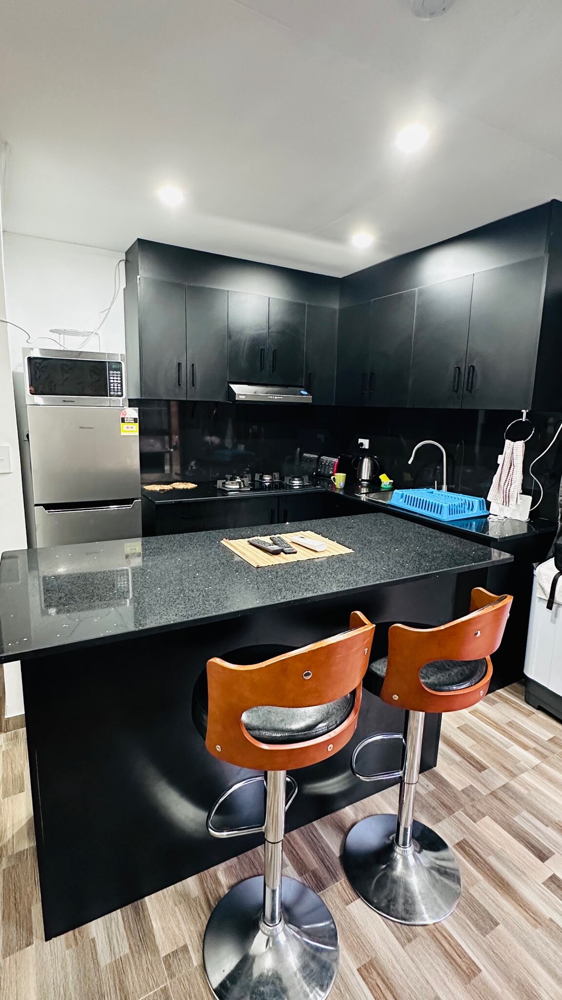 A modern kitchen features sleek black cabinetry and a granite countertop. Two bar stools with wooden seats are positioned at the counter. A stainless steel refrigerator is visible, along with various kitchen appliances and utensils, contributing to a functional cooking space.