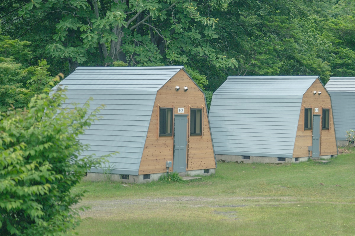 Two cozy cabins are positioned on a grassy area, surrounded by lush greenery. Each cabin features a sloped roof and multiple windows, providing a straightforward design. The doors are centered, leading into the inviting interiors.