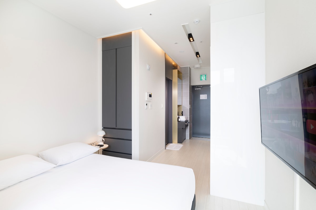 A bright, modern bedroom features a spacious bed with crisp linens, positioned near a wall-mounted television. Soft lighting highlights the sleek cabinetry and minimalist decor. The open layout allows for easy access to the adjacent bathroom and entrance area, contributing to a sense of openness.