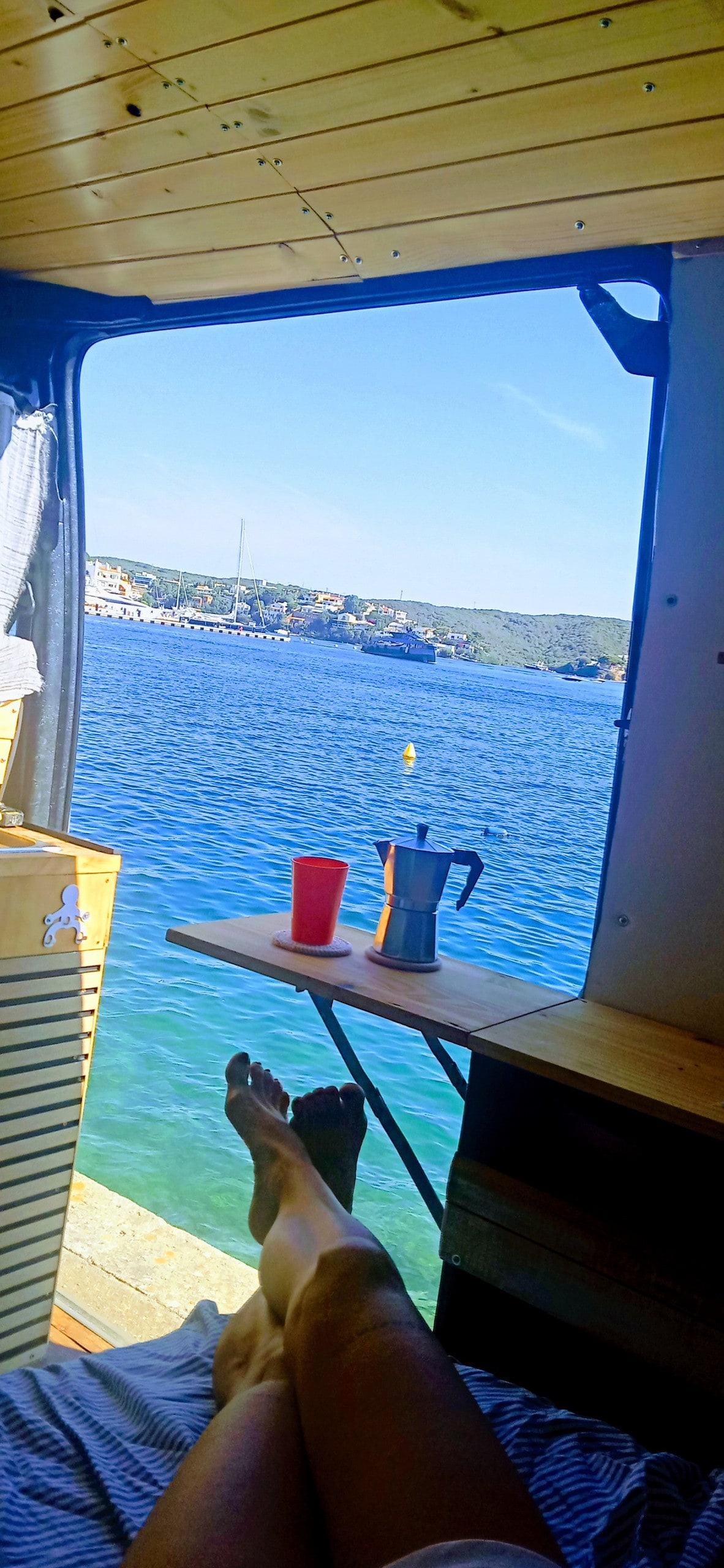 A serene view from within the accommodation reveals feet relaxed against a wooden surface. A colorful cup and a coffee maker are positioned on a small table next to a large opening that frames clear blue waters and distant hills.
