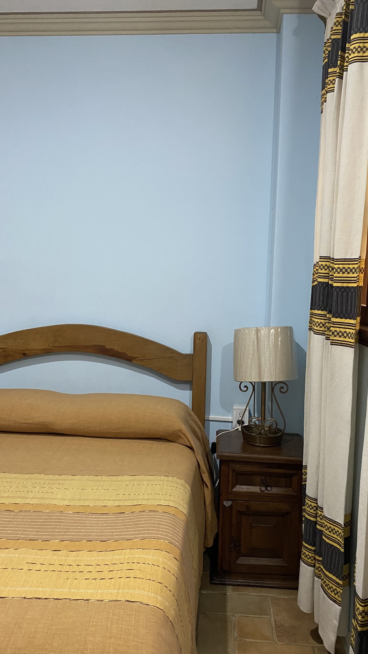 A double room features a wooden bed topped with a beige and yellow striped blanket. A nightstand with a decorative lamp sits beside the bed, and textured curtains frame the window, adding warmth to the overall design.