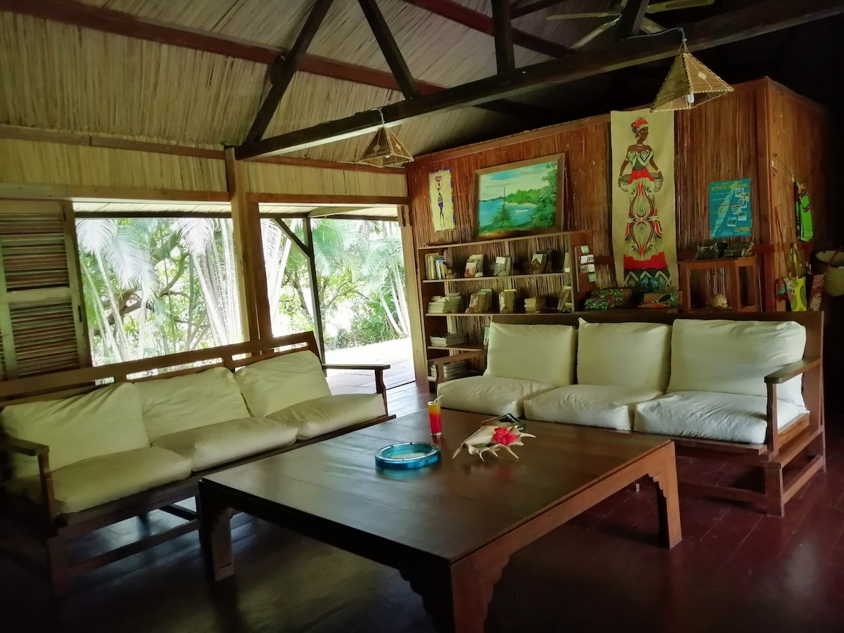 An inviting communal living area features a large wooden table surrounded by comfortable seating. On the walls, local artworks and crafts provide a cultural touch. Large windows offer views of the tropical garden, with natural light enhancing the space's warmth.