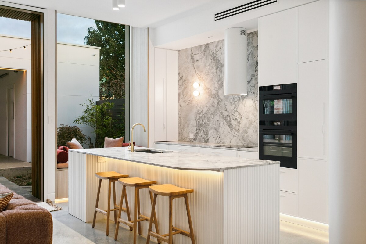 A modern kitchen is highlighted by a spacious island with a marble countertop and three wooden stools. Sleek cabinetry and built-in appliances complement the light-filled space, while warm lighting accents the island's base. Outdoor views are visible through the open doorway.