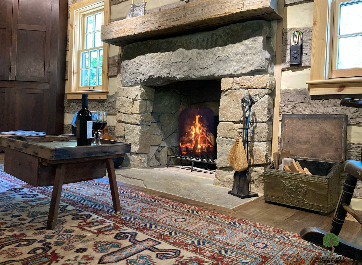 A rustic living room showcases a prominent wood-burning fireplace with a granite stone façade, gently glowing with flames. An antique coffee table sits in front, alongside two glasses of wine. The space features a colorful area rug and wooden accents, enhancing the cabin's charm.