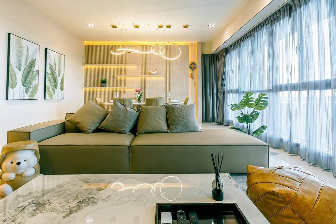 A modern living area features a spacious sectional sofa adorned with plush cushions. Behind, a dining space is illuminated by soft lighting. Large windows allow natural light to brighten the room, while potted greenery adds a touch of nature. Decorative wall art enhances the ambiance.