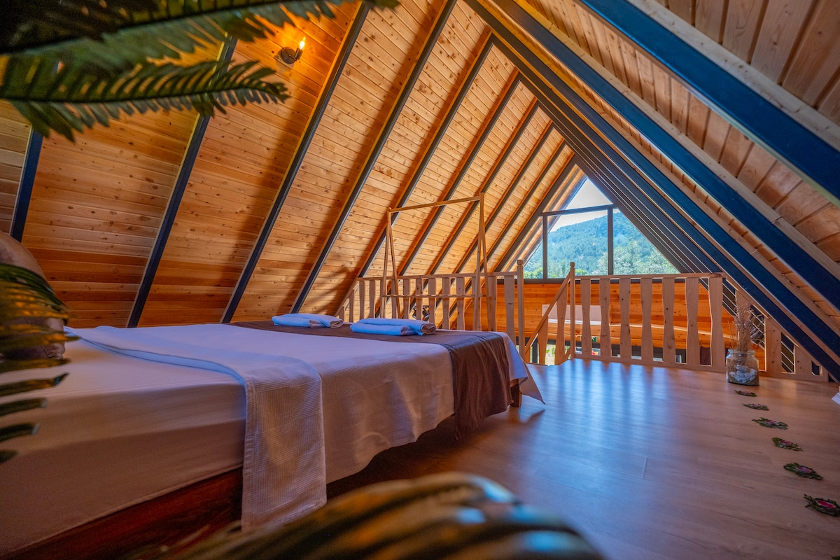 A bright attic space features a spacious bed with neatly arranged linens. Exposed wooden beams and sloped ceilings add character, while natural light floods in through a large window, providing views of the surrounding landscape.