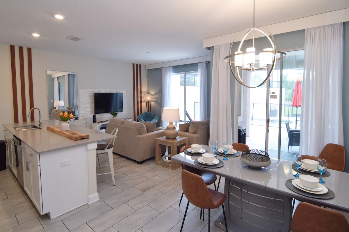 An open-plan living area showcases a modern design, featuring a kitchen with a large island, a dining table set for six, and a cozy lounge space. Large sliding doors provide access to the outdoor pool area, with natural light illuminating the room through sheer curtains.