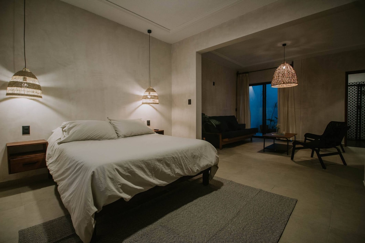 The spacious bedroom features a large bed with crisp white linens. Soft light from woven pendant lamps creates a serene ambiance. A seating area with a sofa and a coffee table is visible, providing a comfortable space for relaxation. A large window allows natural light to enter.