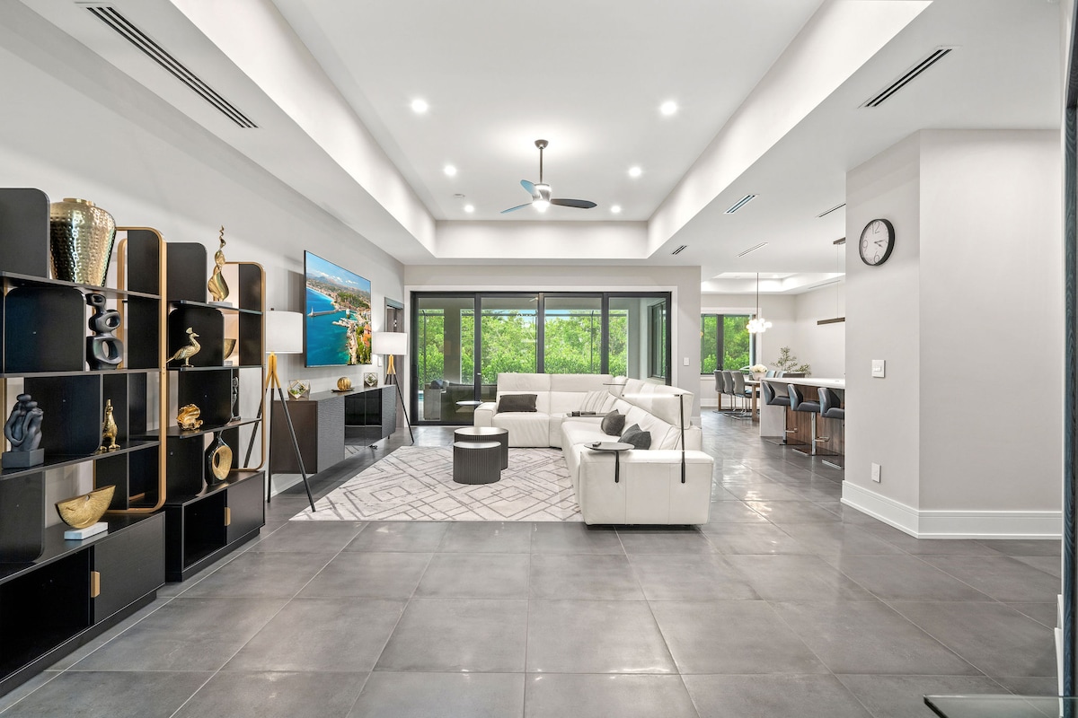 A modern living area features a sectional sofa and an 85-inch smart TV against a minimalist backdrop. Large windows offer views of the outdoors, while an open layout connects to the dining area. Decorative shelving units add a touch of style to the space.