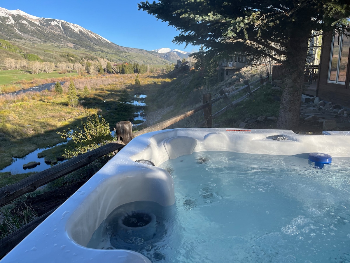 A hot tub is situated on a wooden deck, offering a clear view of the East River and surrounding mountains. The water's surface is gently rippled, reflecting the blue sky above, while trees line the banks of the river, enhancing the natural landscape.