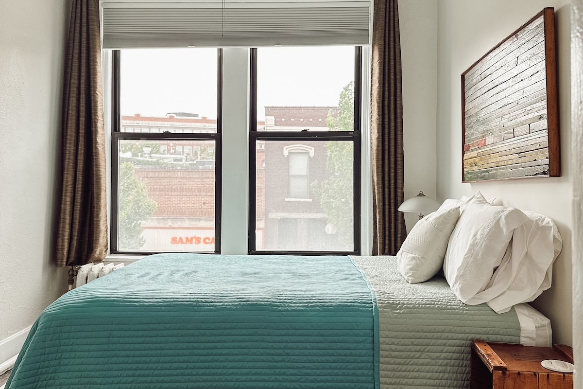A bright bedroom features a spacious bed with a textured aqua quilt and an assortment of white pillows. Two large windows provide natural light and a view of the street, while light-colored curtains frame the windows. A piece of rustic wall art adds visual interest.