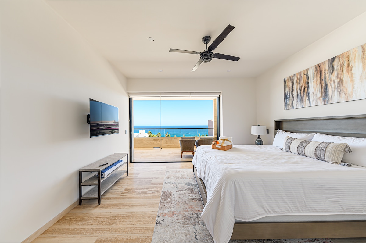 A spacious bedroom is equipped with a king-size bed dressed in crisp white linens. A wall-mounted television is mounted across from the bed. A private balcony is visible through glass doors, offering views of the ocean. A ceiling fan adds comfort to the airy space.