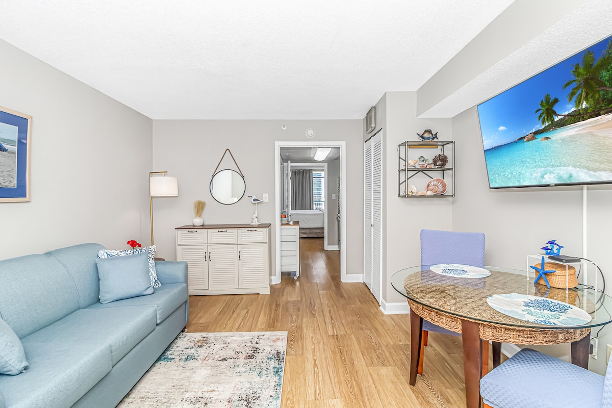 An updated living space features a light blue sofa adorned with decorative pillows. A round dining table with two blue chairs is positioned nearby. A wall-mounted television displays a tropical scene, while coastal-themed artwork decorates the walls. Natural light fills the area, enhancing the warm wood floors.