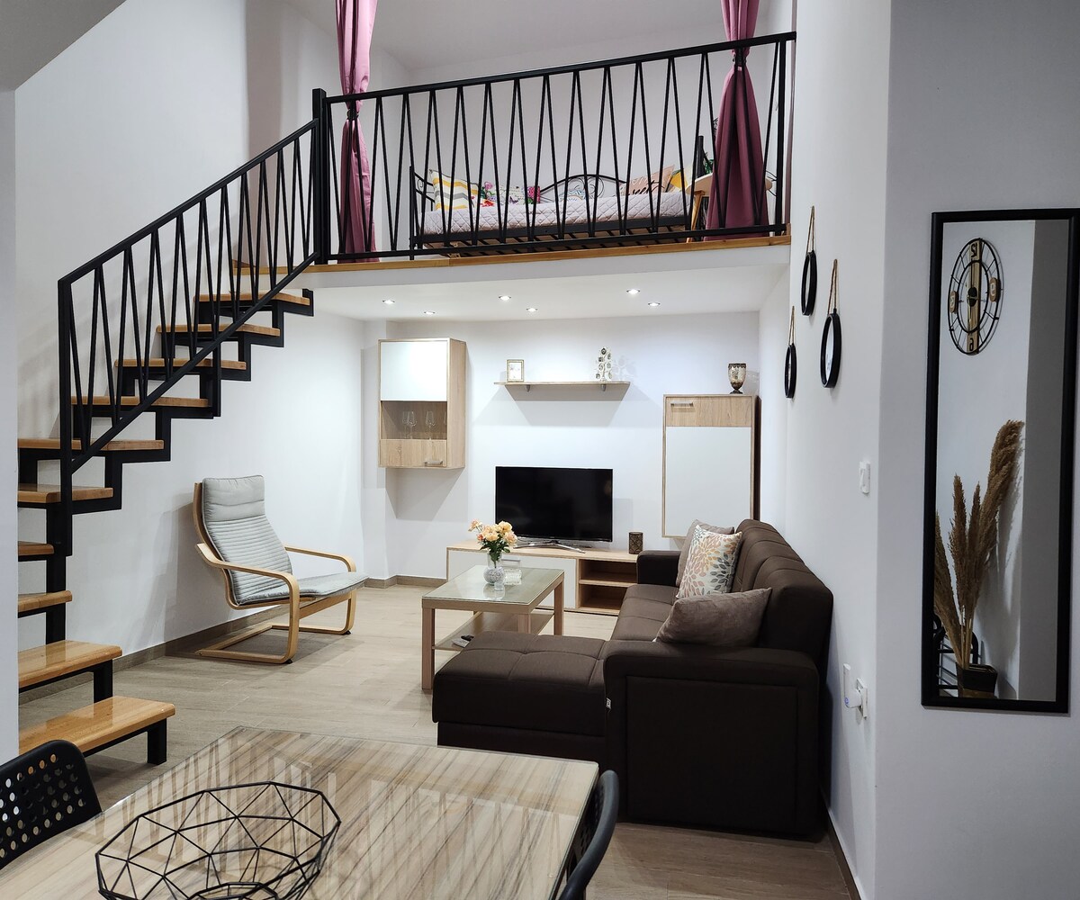 A modern loft apartment features a spacious living area with a comfortable sofa and a glass coffee table. A staircase with wooden steps leads to a cozy sleeping loft adorned with colorful bedding. Soft lighting from recessed fixtures enhances the welcoming ambiance.