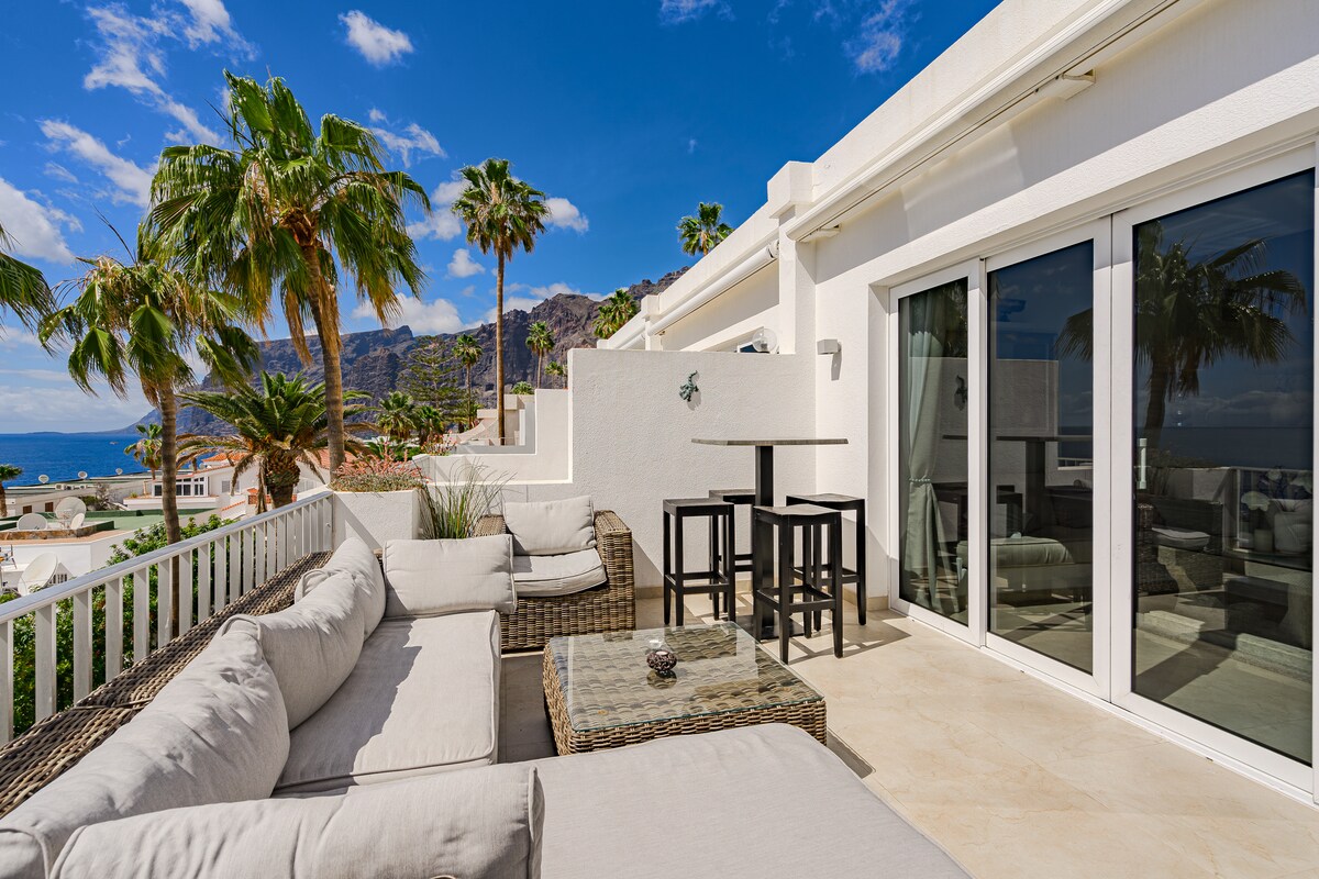 An outdoor terrace is seen with a modular seating arrangement and a small dining table. Tropical palm trees and distant ocean views are visible, creating a relaxing environment. The area is well-lit, highlighting the neutral tones of the furniture against the bright sky.