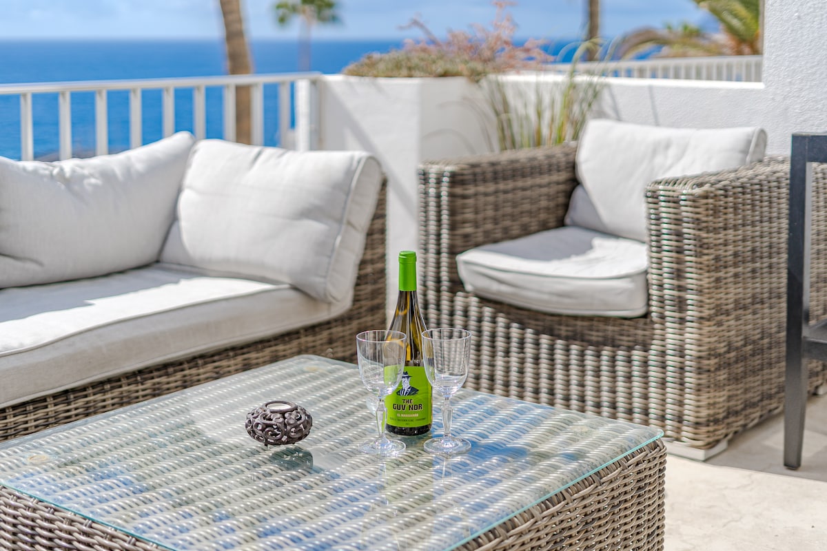 A terrace seating area is presented with a glass-top table, two comfortable cushioned armchairs, and a bottle of wine accompanied by two glasses. The ocean view is visible in the background, framed by greenery and palm trees.