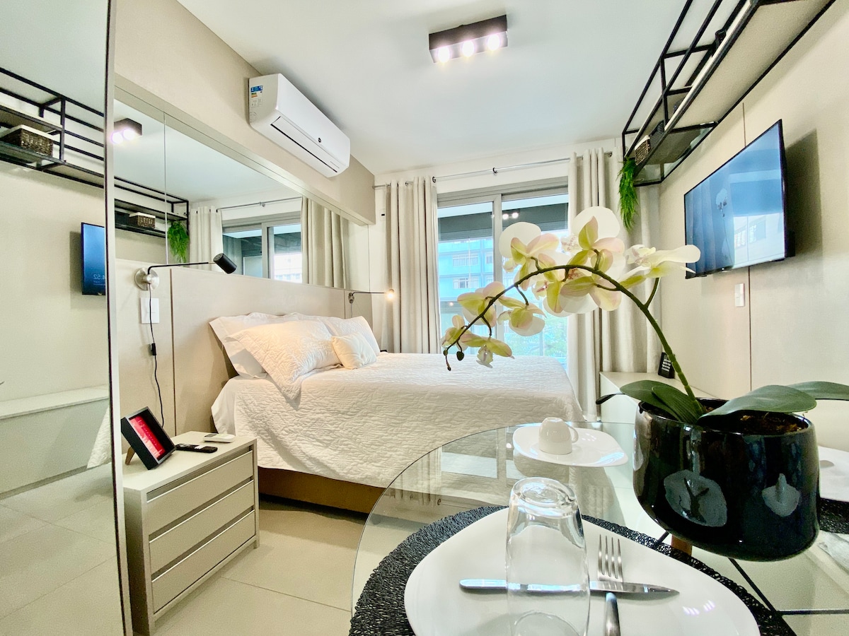 A modern studio space showcases a spacious bed with a textured white coverlet. A glass table is set with simple dining essentials. Natural light filters through large windows, and an artificial orchid adds a touch of greenery. Wall-mounted TVs are visible for entertainment.