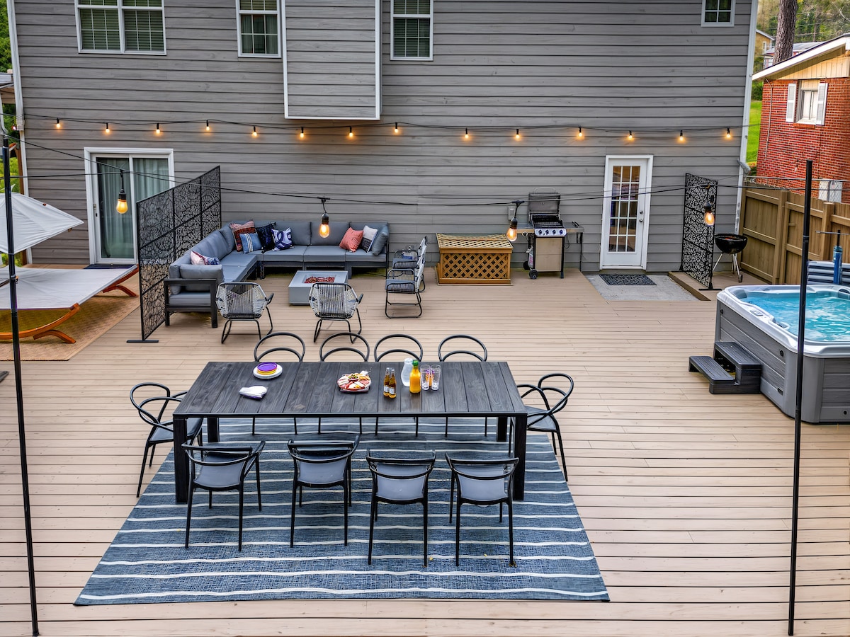 An expansive outdoor patio features a large dining table with seating for ten, surrounded by stylish chairs. A comfortable sectional sofa offers additional seating, while a hot tub and grill are positioned at the patio's edge. String lights hang overhead, creating a warm atmosphere.