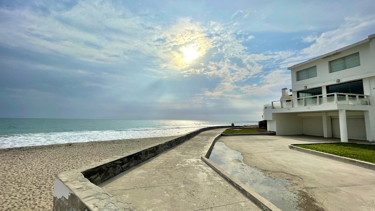 A coastal view is presented, showcasing a smooth pathway leading to the beach. The ocean reflects soft sunlight under a cloudy sky, while a modern house stands nearby with a balcony overlooking the water. Sandy shores stretch into the horizon.