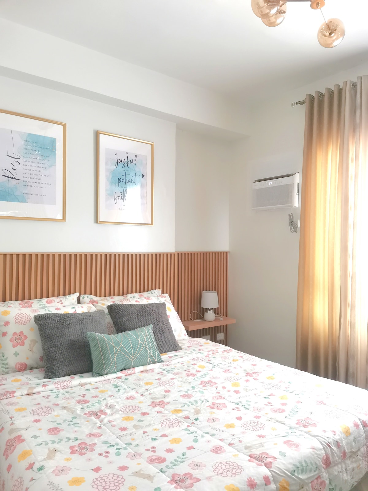 A bright bedroom features a floral patterned bedspread draped over a spacious bed, complemented by decorative pillows. Two framed art pieces adorn the wall above the headboard, while a window with sheer curtains allows natural light to filter in, showcasing a calming atmosphere.