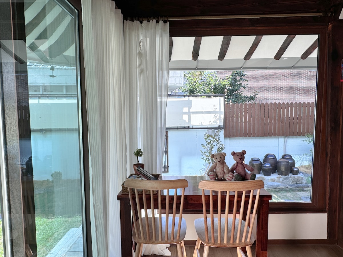 A charming seating area features two wooden chairs positioned at a table, adorned with soft toys. Large glass windows frame the serene outdoor view, allowing natural light to flood the space. Sheer white curtains provide a gentle touch, enhancing the tranquil ambiance.