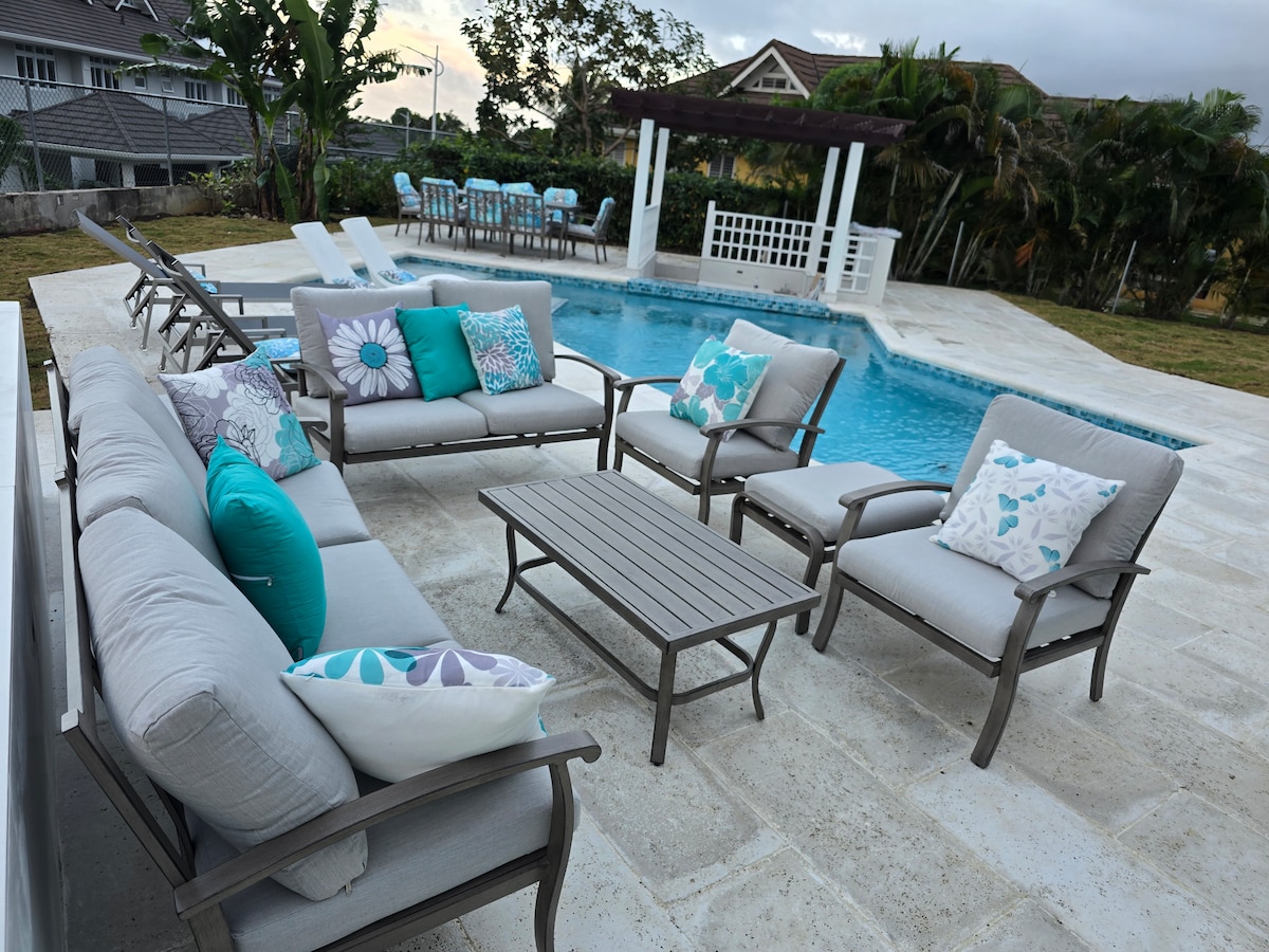 An outdoor seating area features a set of comfortable couches and chairs arranged around a low table, adorned with decorative cushions in shades of blue and green. Behind the seating, a private pool glimmers under the sun, surrounded by a spacious deck.