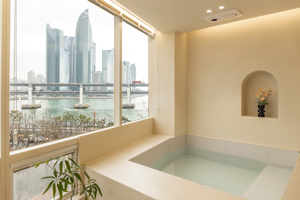 A modern indoor jacuzzi is positioned by large windows, offering a view of the city skyline and the bridge over the water. Minimalist decor includes a small potted plant on the ledge, complementing the serene atmosphere.