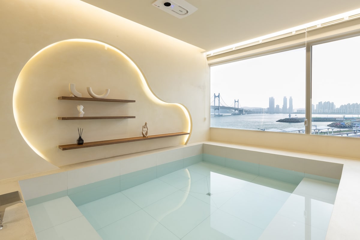 A serene indoor jacuzzi is positioned next to a large window, offering views of the Gwangan Bridge and the coastline. Soft, warm lighting highlights the modern design, while minimalist shelves with decorative items enhance the calming atmosphere of the space.