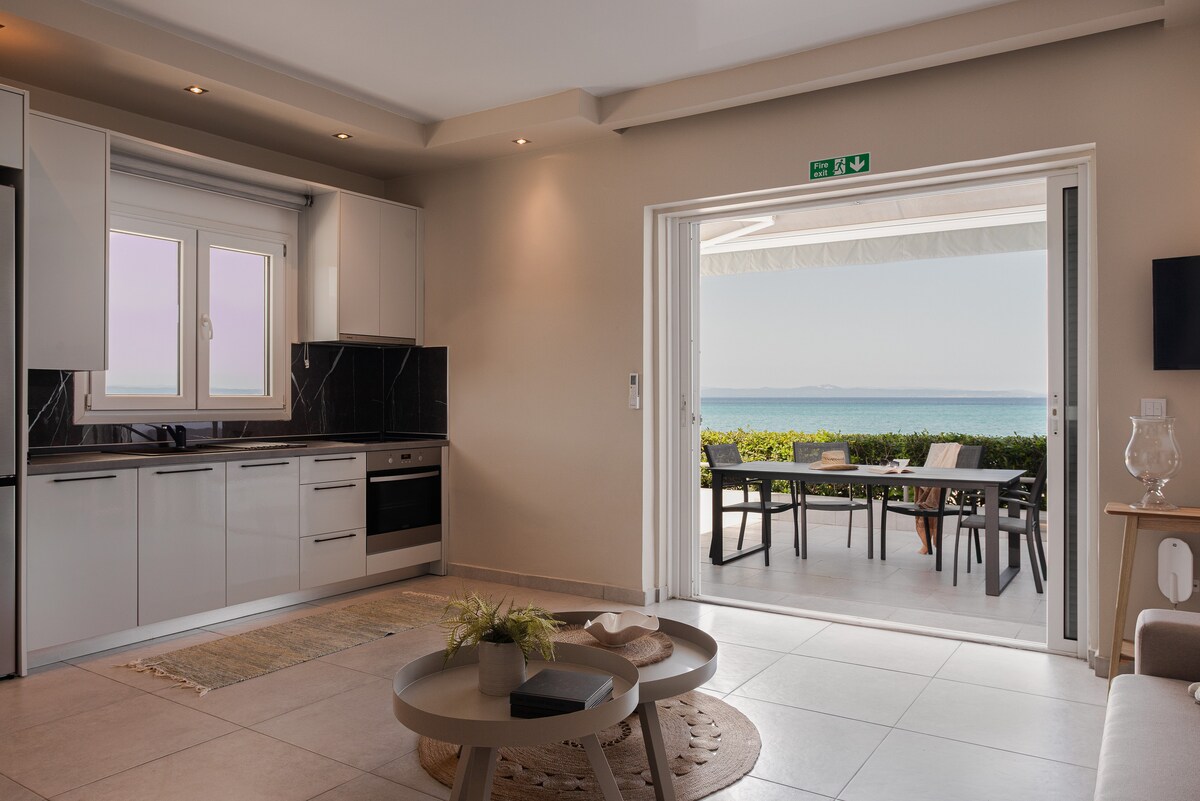 A modern kitchen is visible, featuring sleek white cabinets and black countertops. An open doorway leads to an outdoor dining area with a table and chairs, framed by views of the sea. Natural light fills the space from the large windows.