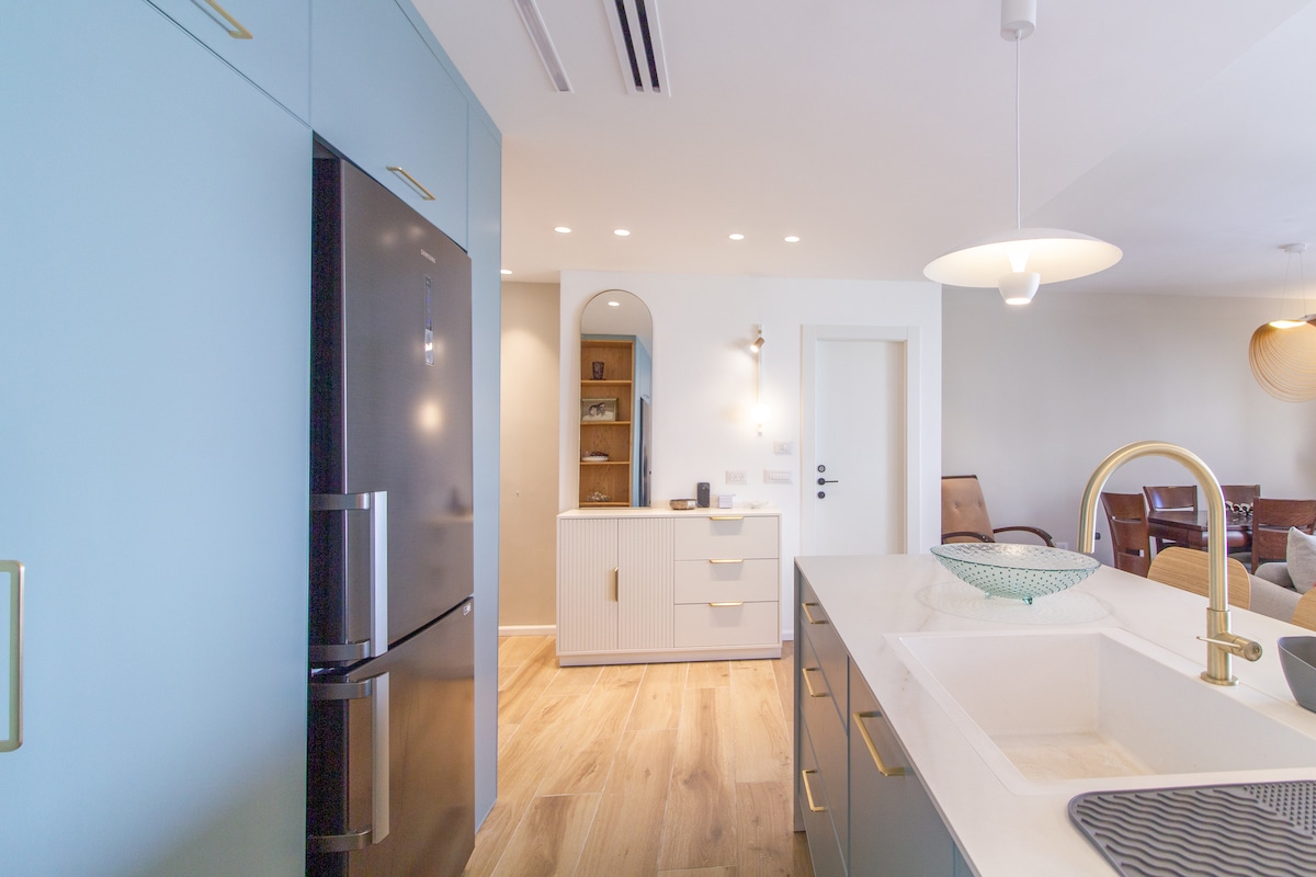 The spacious kitchen area features modern cabinetry in a light blue tone, complemented by a sleek black refrigerator. A large island with a sink is at the forefront, while natural light illuminates the open layout, showcasing an inviting connecting living space.