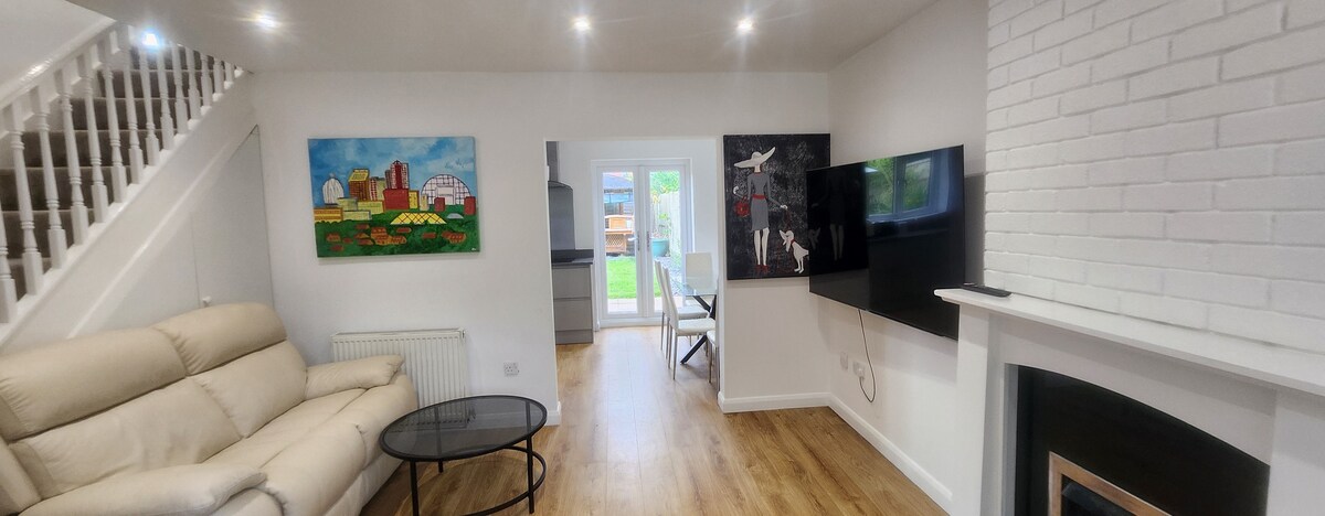 A light-filled lounge area features a cream reclining sofa, a glass coffee table, and a mounted flat-screen TV. Artwork adorns the walls, complementing the contemporary decor. A set of glass doors leads to the garden, extending the living space outdoors.