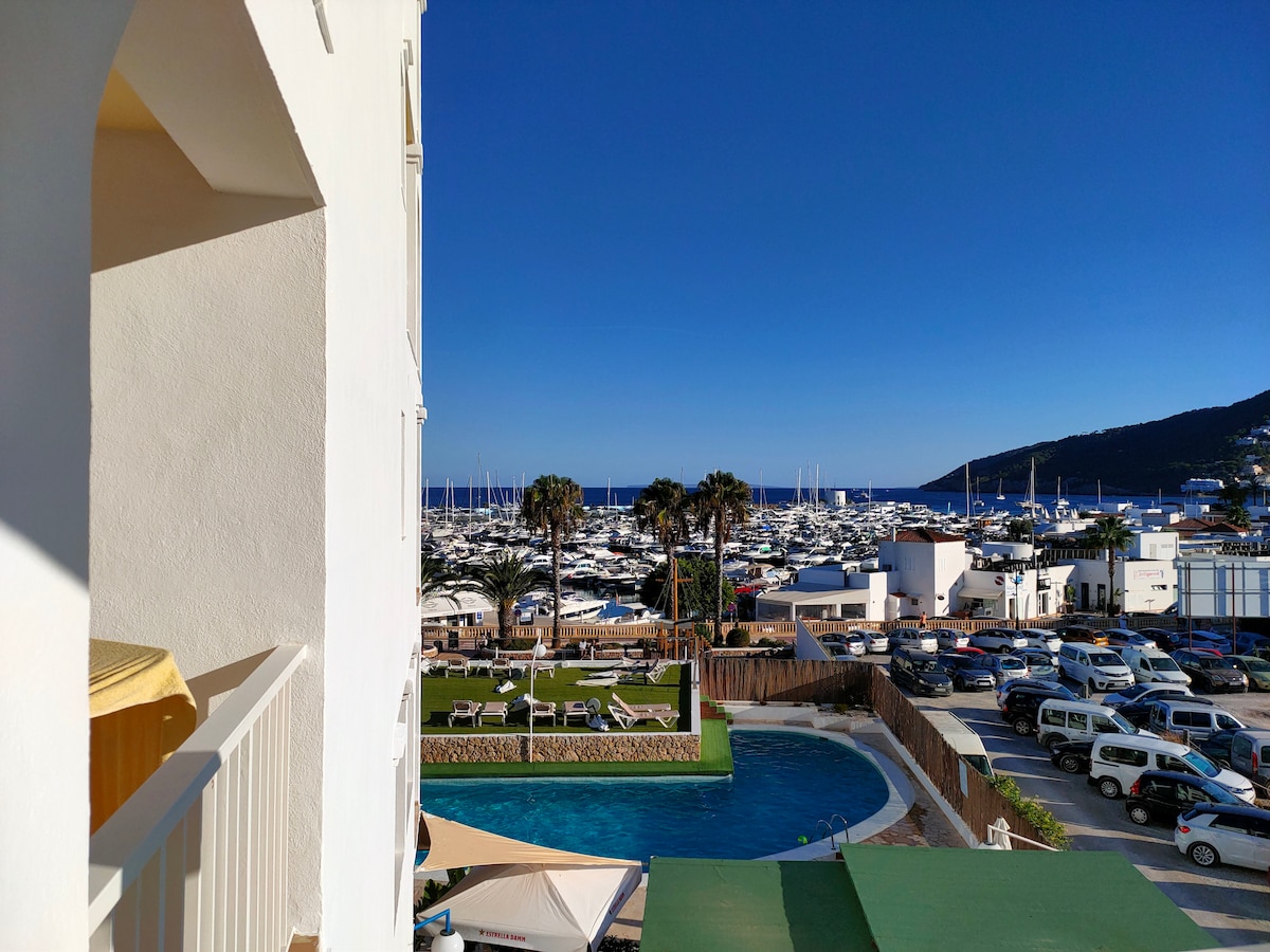 A view from a balcony captures the marina filled with boats and yachts, framed by palm trees. Below, an outdoor pool is visible alongside a landscaped area with lounge chairs. A parking area and additional structures are observed in the foreground.