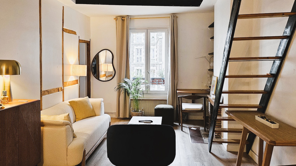 The living area features a spacious white sofa with a yellow accent pillow, complemented by a wooden coffee table. A small desk is positioned beside a window with curtains, bringing natural light into the room. A wooden ladder leads to a mezzanine area.