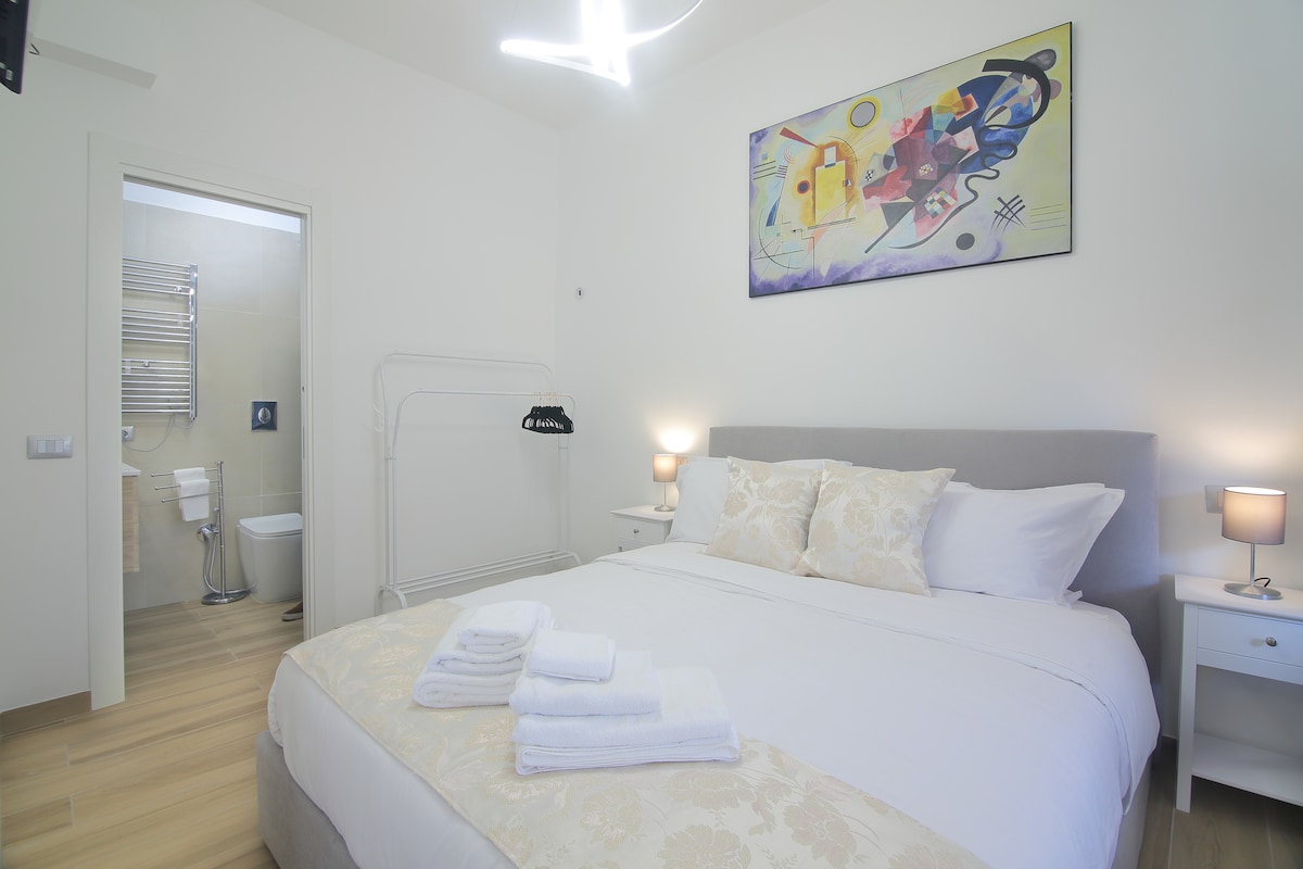 A well-lit bedroom features a spacious bed adorned with decorative pillows and neatly arranged towels. A colorful artwork is displayed above the bed, while an ensuite bathroom with modern fixtures is visible through a doorway. Soft lighting from bedside lamps enhances the serene ambiance.