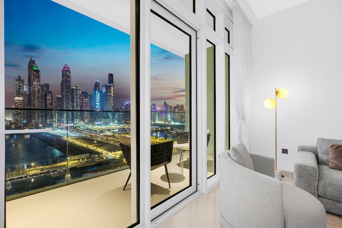 A modern living space features expansive floor-to-ceiling windows, offering stunning views of the city skyline and waterfront. The balcony showcases stylish outdoor seating, while warm indoor lighting creates a cozy atmosphere. A comfortable sofa completes the elegant interior.