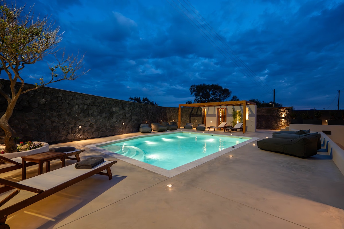 A serene outdoor area at twilight showcases a sparkling swimming pool surrounded by comfortable loungers and soft lighting. Stone walls provide privacy, while a tree adds greenery to the scene. The inviting pool area is ideal for relaxation and enjoying peaceful moments.