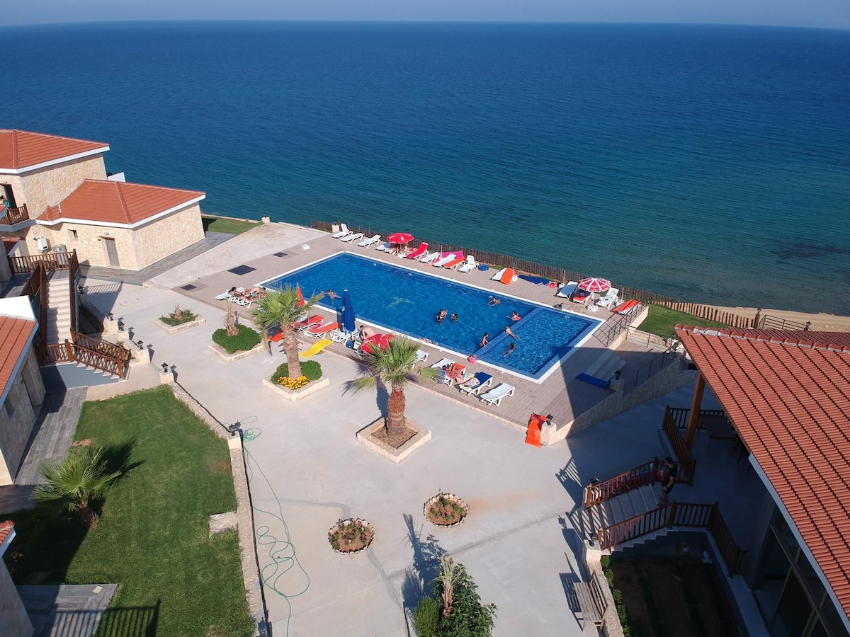 A large swimming pool is surrounded by lounge chairs and umbrellas, offering a space for relaxation. The pool area is bordered by well-maintained landscaping, featuring palm trees. The sea is visible in the background, providing a serene view of the horizon.