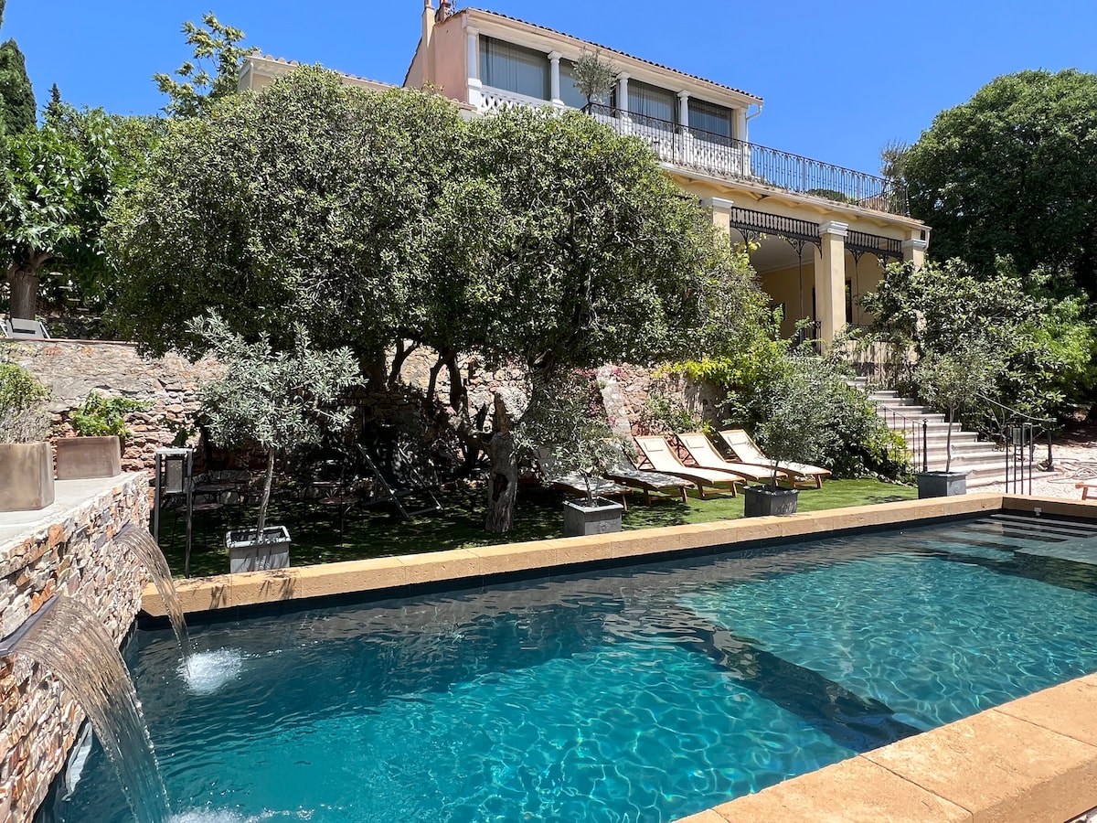 A sparkling pool is bordered by a stone wall, with two cascades creating gentle waterfalls. Eight sun loungers are arranged under the shade of trees, while a lush garden surrounds the area. The elegant house is visible in the background, featuring outdoor terraces.