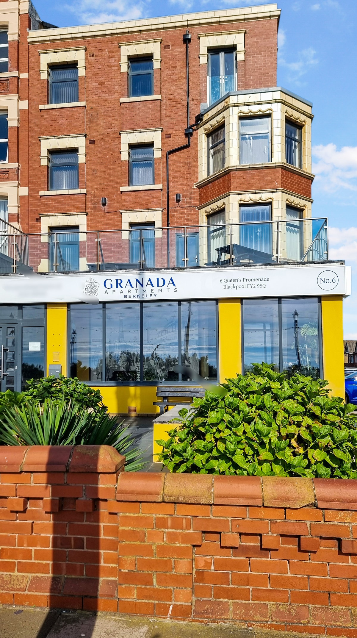 A modern building features large glass windows and a vibrant yellow entrance. The structure showcases a mix of red brick and stone detailing. Lush greenery is planted in front, providing a natural contrast to the urban setting. A clear sky is visible in the background.