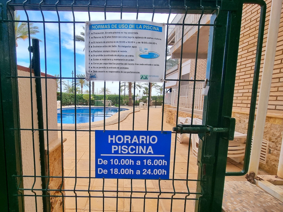 A gated area leads to the community pool, featuring clear water and lounge chairs nearby. Signs display pool usage rules and opening hours, offering guidance for guests. Surrounding greenery and palm trees create a relaxed outdoor environment.