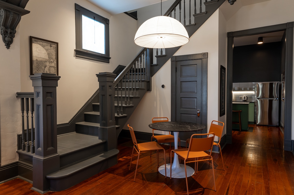 An inviting dining area features a round black table surrounded by four orange chairs. The dark staircase, with detailed railings and a window above, adds a striking contrast to the hardwood floors. Stainless steel appliances are visible in the kitchen area through an open doorway.