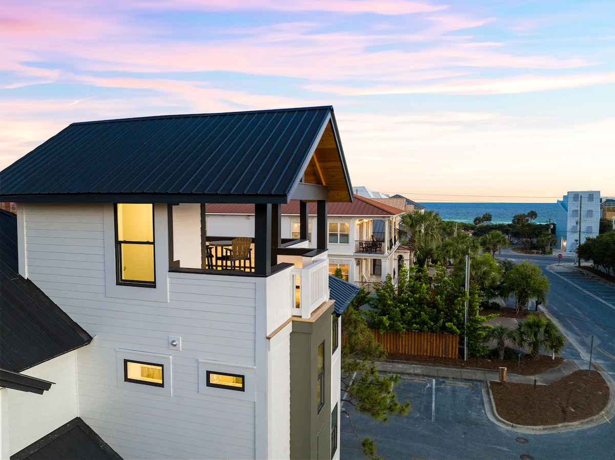 The exterior view captures the top corner of the newly constructed home, highlighting a covered balcony with seating. The ocean can be seen in the distance, framed by nearby palm trees and residential buildings, suggesting a serene coastal setting.