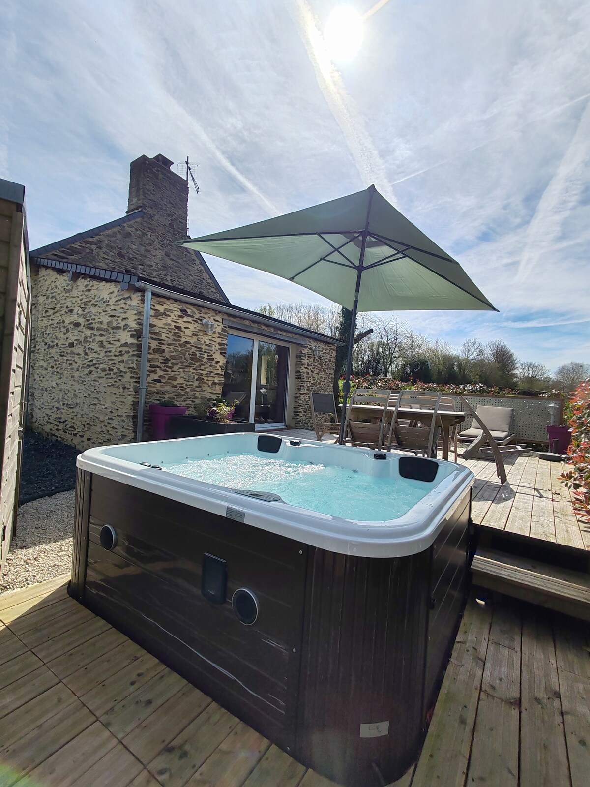 A spacious wooden terrace is complemented by a jacuzzi, inviting relaxation. An umbrella provides shade over the seating area, which includes a table and chairs. Sunlight reflects off the water, creating a calming atmosphere, while the rustic stone house serves as a backdrop.