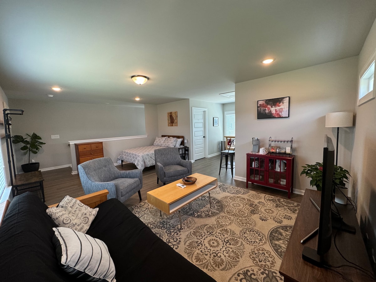 A spacious living area is arranged with a sofa and two armchairs. A wooden coffee table is centered on a decorative area rug. A flat-screen TV is positioned on a red console table. In the background, a bed and a second sleeping area are visible.