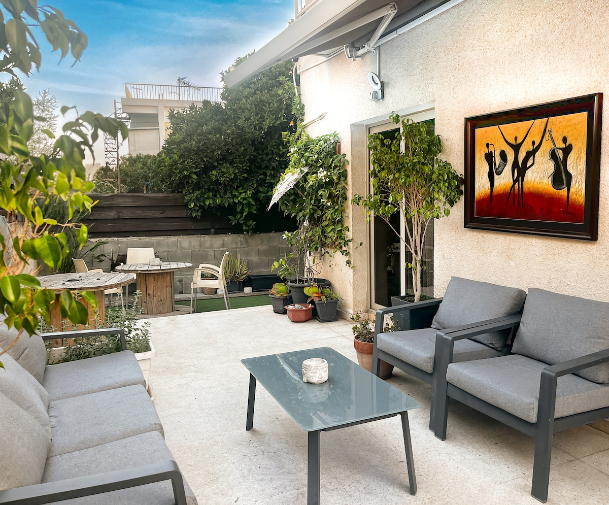 An outdoor seating area features comfortable gray lounge chairs and a glass table, surrounded by lush greenery and potted plants. A colorful piece of art is displayed on the wall, adding vibrancy to the serene space. Natural light enhances the inviting atmosphere.