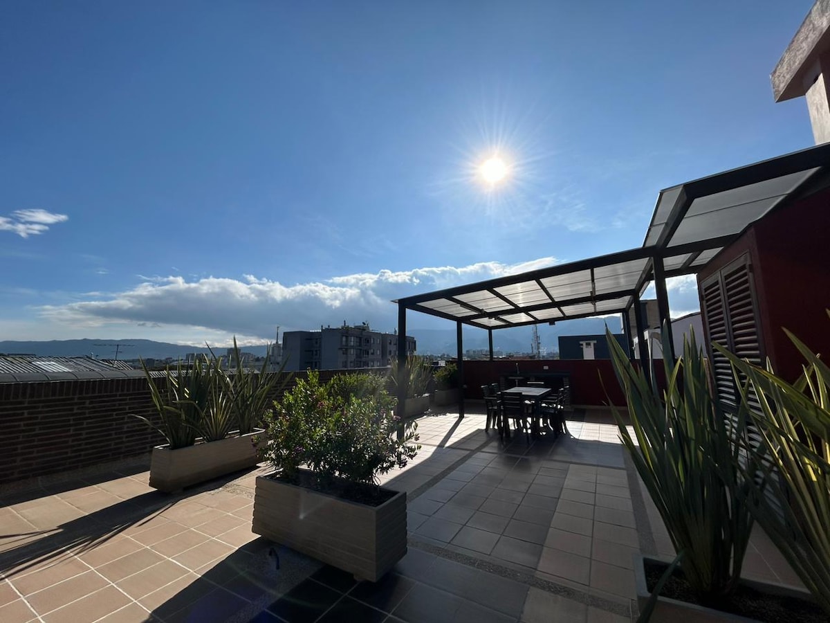 A spacious rooftop terrace is bathed in sunlight, featuring a pergola and outdoor seating area with a dining table. Planters with green plants adorn the space, creating a relaxing environment for guests. The horizon showcases distant buildings against a clear blue sky.