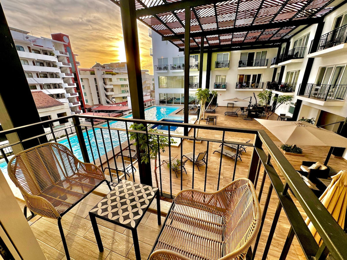 A serene balcony view showcases two wicker chairs and a small table overlooking a vibrant pool area. The sunset casts warm hues over the scene, highlighting the surrounding buildings and lush greenery within the courtyard.