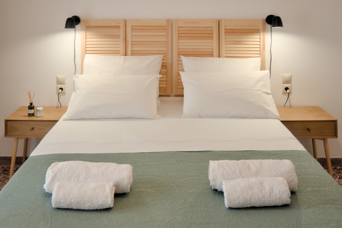 A spacious sleeping area features a double bed with a wooden headboard and crisp white linens. Two matching bedside tables are positioned on either side, each illuminated by wall-mounted lamps. Fresh towels are neatly arranged on the green bedspread, enhancing the room's organized appearance.